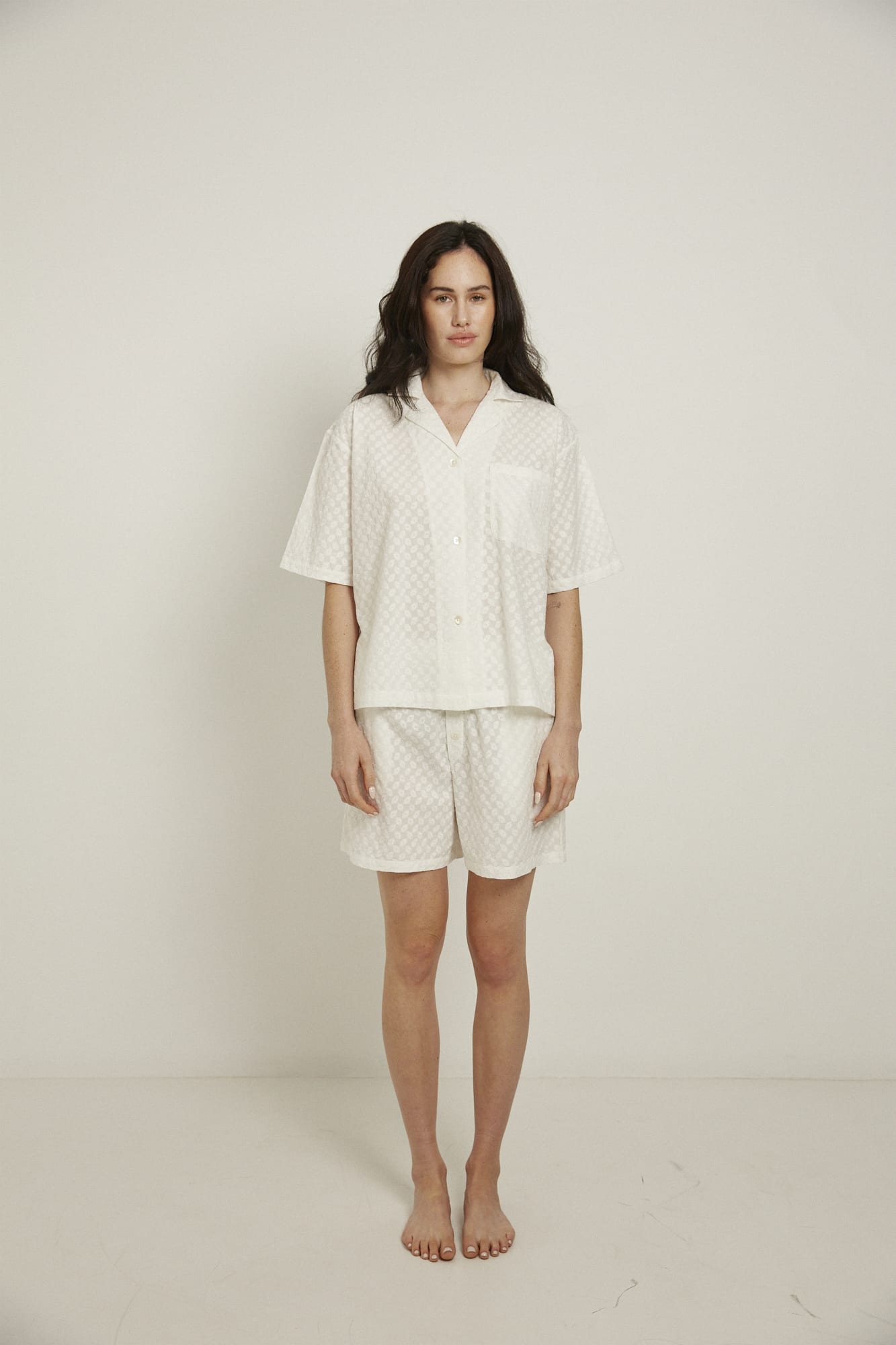 Women’s pyjama set including a short sleeve shirt and shorts. Made from 100% organic cotton, in white colour featuring delicate all over embroidery. The shirt has shell buttons, a front pocket and a back pleat with locker loop detail.  The shorts feature natural shell buttons and an elasticated waistband for comfort.